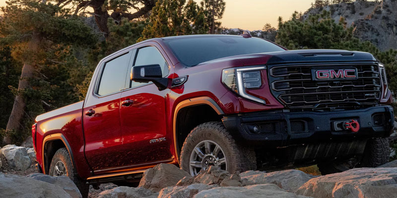A red GMC pickup truck parked on a rocky trail in a forested, mountainous area.