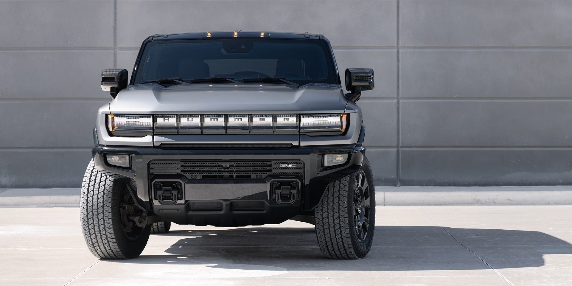 Front view of a matte grey GMC Hummer EV truck parked against a concrete wall.